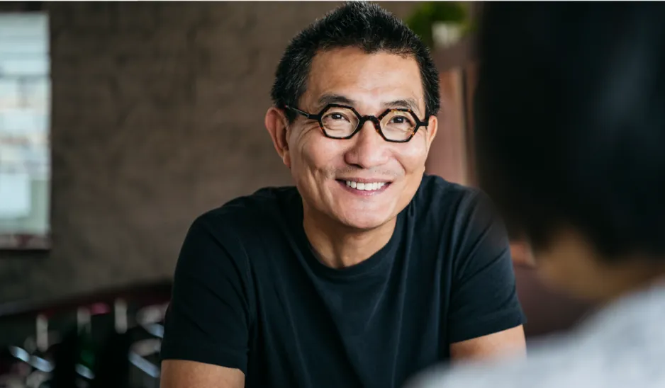 The man wearing glasses smiling as he converses, with a window in the background