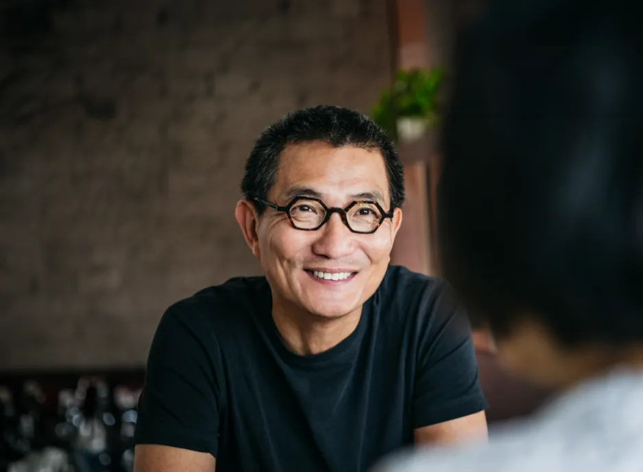 The man wearing glasses smiling as he converses, with an extended brick wall background at the top.