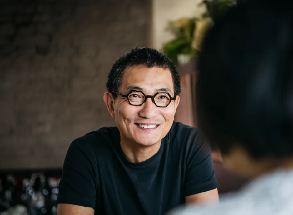 The man wearing glasses smiling as he converses, with an extended brick and plaster wall background at the top.