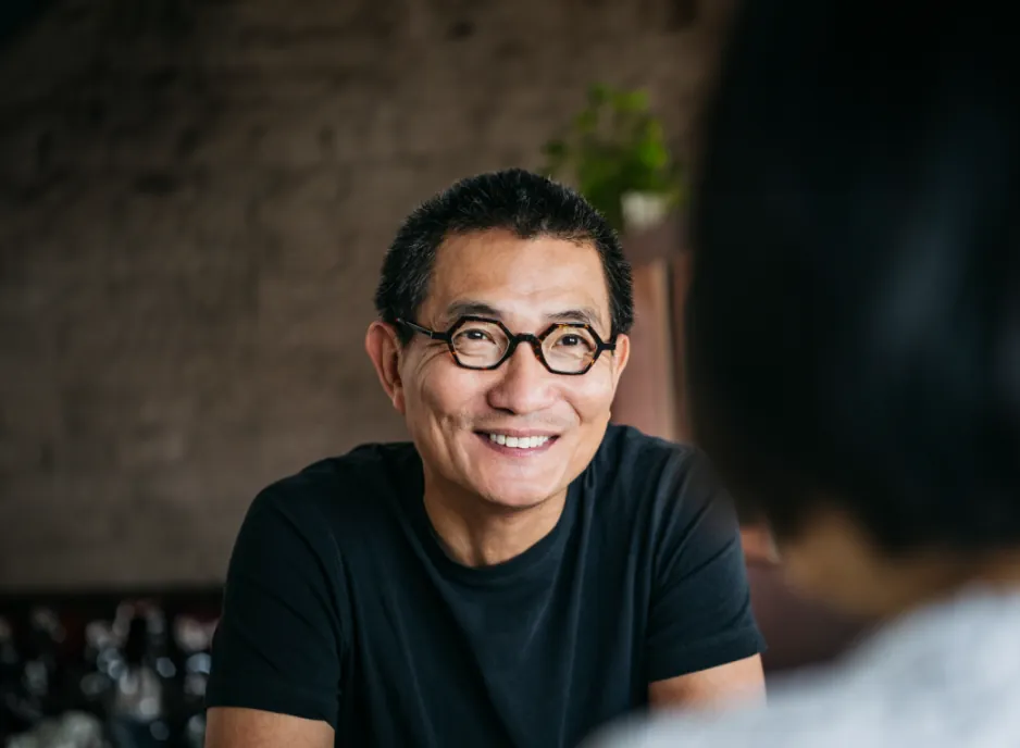 The man wearing glasses smiling as he converses, with a solid brick wall in the background