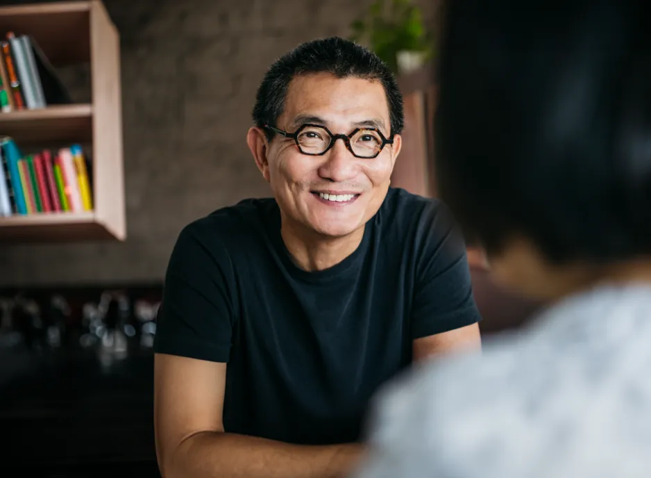 The man wearing glasses smiling as he converses, with a bookshelf in the background that has incorrect perspective