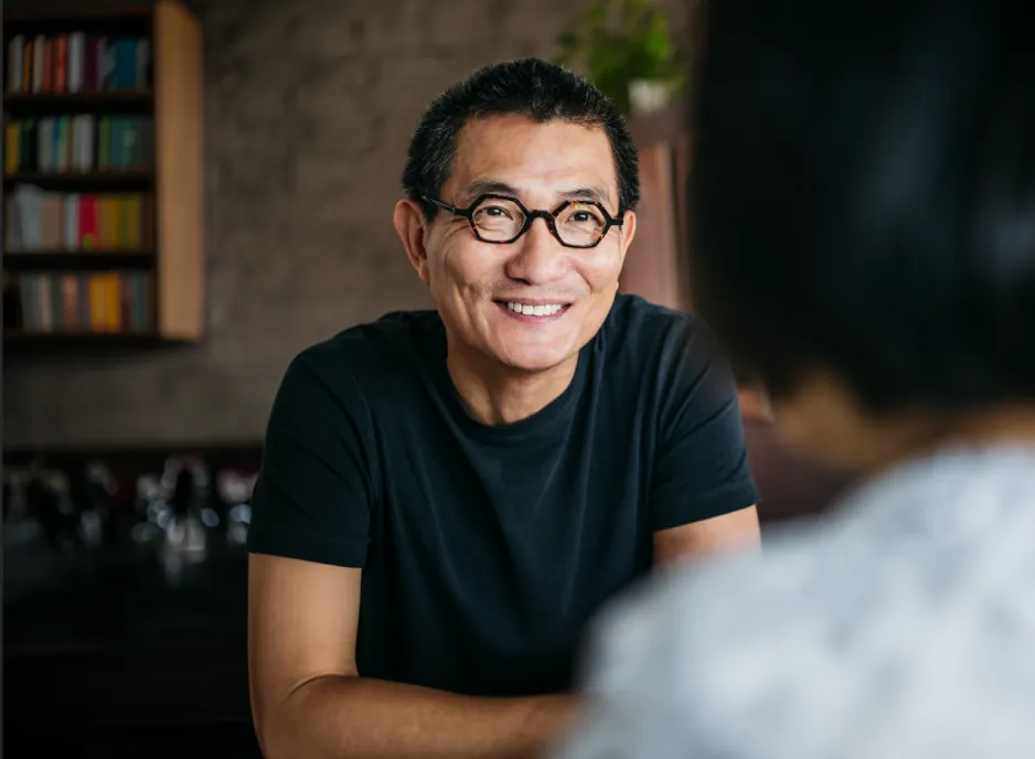 The man wearing glasses smiling as he converses, with a solid brick wall in the background