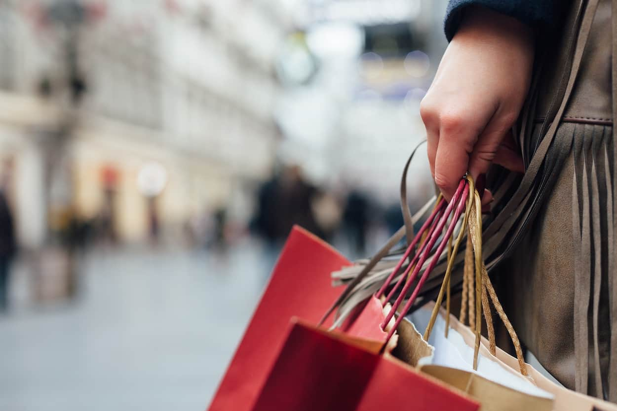 Closeup of a person's hands, holding several shopping bags, with a busy plaza in the background.