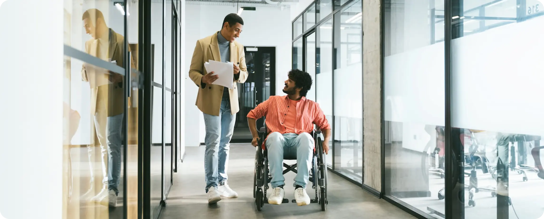 A person in a wheelchair speaking with a friend in an office hallway.
